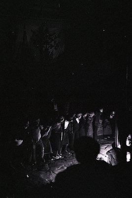 Black-and-white nighttime gathering in a forest clearing, featuring a semi-circle of seated and standing figures. Light sourc...