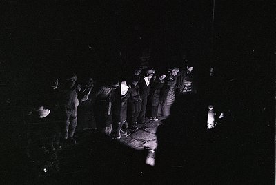 Black-and-white nighttime gathering of 12+ individuals in a dimly lit outdoor setting, illuminated by a single lantern. Group...