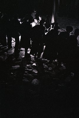 Silhouetted crowd gathered around a central fire in a dimly lit, cobblestone plaza. Silhouettes suggest evening or nighttime ...