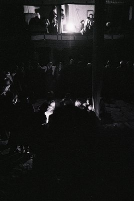 Vintage black-and-white shot of a dimly lit indoor gathering, likely a 1950s–1960s social event. Crowded room with silhouette...
