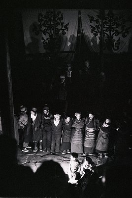 Traditional folk ensemble performing in dimly lit indoor setting, likely a rural or cultural hall. Group of 10+ individuals i...