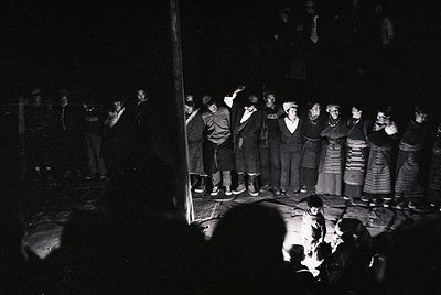 Black-and-white photo of a traditional folk choir performance in a dimly lit hall, likely Eastern European. Women in striped ...