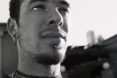 Close-up portrait of a person with facial piercings (nose, ear, lip) and a goatee, captured in high-contrast black-and-white....
