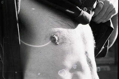 Close-up of a male torso featuring a nipple piercing with a small circular stud. High-contrast black-and-white composition hi...