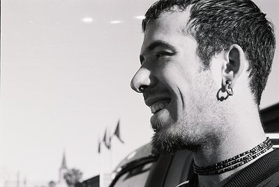 Portrait of a bearded man with a nose ring and earring, smiling warmly. He wears layered necklaces and a dark top. Background...