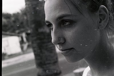 Close-up black-and-white portrait of a person with a nose ring, captured through a rain-streaked window. Reflections blur out...