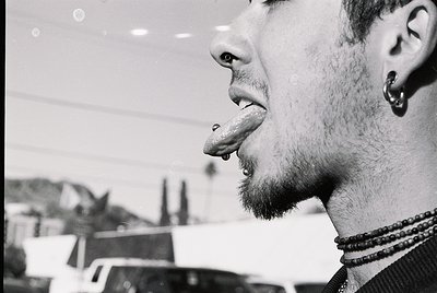 Close-up of a person with a tongue piercing and facial piercings, set against an urban indoor background. Black-and-white pho...