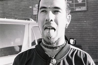 Black-and-white portrait of a person with tongue piercing, nose ring, and layered necklaces, set against an urban backdrop. S...