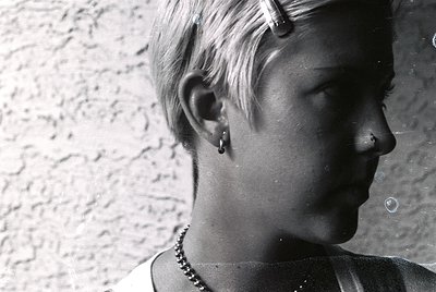 Close-up of a person with short, textured hair and minimalist jewelry—earrings and a beaded necklace—against a rough-textured...