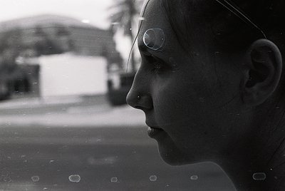 Close-up black-and-white portrait of a person gazing upward, captured through a rain-spotted window. Urban background shows b...