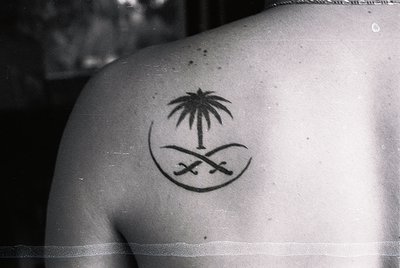 Black-and-white tattoo of Saudi Arabian national emblem—palm tree, crescent moon, and two crossed swords—on upper back. Symbo...