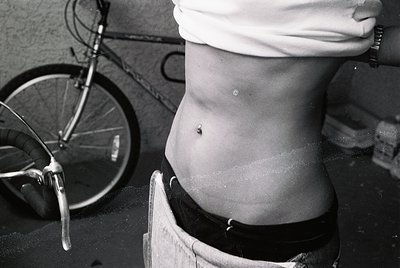 Black-and-white close-up of a person’s torso seated on a wooden barrel, wearing a white headscarf and dark waistband. A bicyc...
