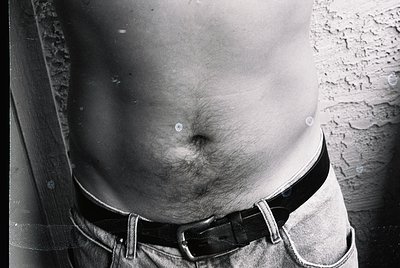 Close-up of a torso in mid-century casual wear, featuring a belted waistband and faded denim. Textured concrete wall in backg...