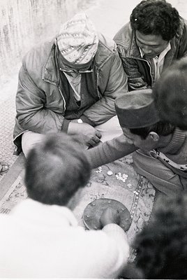 Black-and-white street scene from the 1960s–70s, likely Eastern Europe. Three men in winter coats and headwear gather around ...