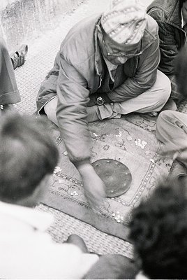 Mid-20th century black-and-white photo of a man in a white cap and glasses meticulously sorting small metal discs on a patter...
