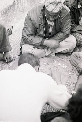 Mid-20th century street scene: Elderly man in traditional headwear and loose trousers mends shoes on a patterned rug, seated ...
