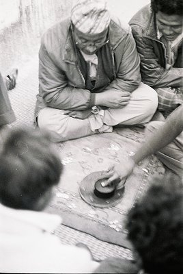 Men seated around a large, folded map in an outdoor setting, likely mid-20th century. Foreground shows a watch and hands adju...