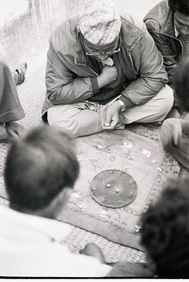 A man in 1960s-era military uniform repairs a shoe using a cobbler’s tool on a patterned floor. Blurred figures in the backgr...