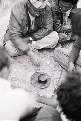 Black-and-white photo of three men playing a traditional board game on a woven mat, likely in a rural setting. Central figure...