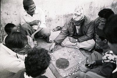 Group of men seated on woven mats in a dimly lit, textured interior, playing a traditional board game. Central figure wears a...