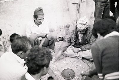 Black-and-white photo capturing a traditional Nepalese ritual or gathering. Elderly man in turban seated on floor, surrounded...