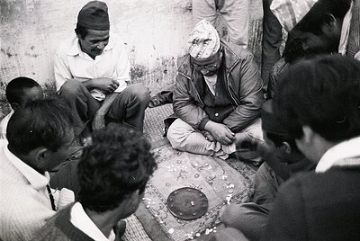 A group of men in traditional attire gathers indoors, seated on a woven mat around a central table. One man, wearing a headsc...