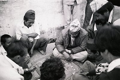 A group of men in traditional attire gathers indoors, seated on a tiled floor around a central figure wearing a headscarf and...