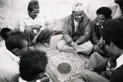 Group of men seated on floor playing traditional board game, likely in South Asia. Mid-20th century attire (1950s–1970s) with...