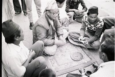 Group of men seated on patterned rugs in traditional attire, engaged in a communal activity—likely a card game or discussion—...