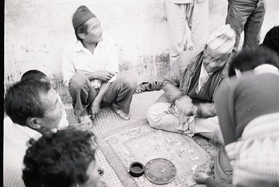 Group of men seated on patterned rugs in a traditional indoor setting, likely a home or communal space. One man wears a turba...