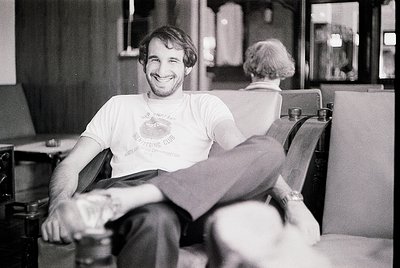 Mid-century barber shop interior with a relaxed man in a vintage "Every Living Thing" T-shirt, seated in a classic upholstere...
