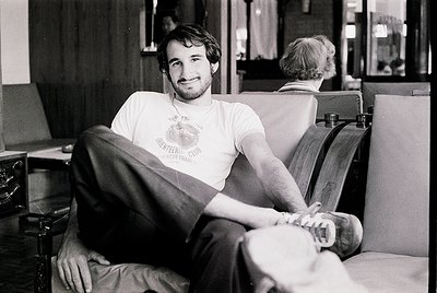 Black-and-white portrait of a relaxed man in a 1970s-style T-shirt ("Keep Peace & Quiet" with a peace symbol) lounging on a l...