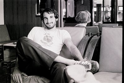 Relaxed man in a 1970s-style T-shirt (with "Greenwich Village" and "Scout Club" text) lounges on a leather armchair in an ind...