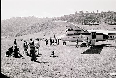 A group of people gathers around a Royal Nepal Airlines floatplane (registration 9N-ABX) on a grassy airstrip, likely in the ...