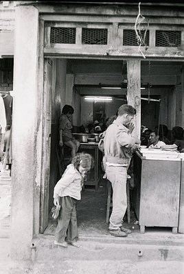 Vintage street-side café or snack bar interior, likely mid-20th century. A man in a light apron serves food from a counter, w...
