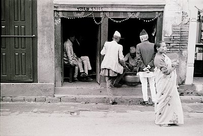 Black-and-white street scene in Nepal, likely Kathmandu, mid-20th century. Traditional attire—men in *dhaka* and *topi*, wome...