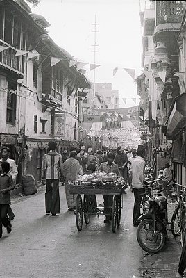 Vibrant street scene featuring a bustling market cart laden with fresh produce, likely melons and vegetables, in a narrow all...