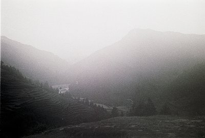 Misty mountain valley with terraced fields and sparse structures, captured in monochrome. Fog obscures distant ridges, emphas...