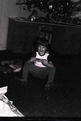 Vintage black-and-white photo of a young boy in a 1970s-style tank top and pants, holding a wrapped gift under a decorated Ch...