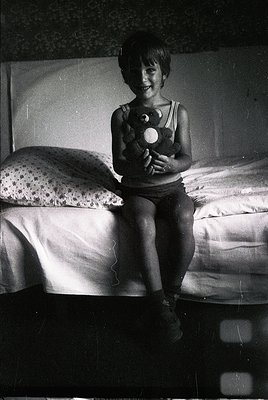A young child sits on a twin bed holding a stuffed toy, likely from the 1950s–1960s. The room features floral-patterned beddi...