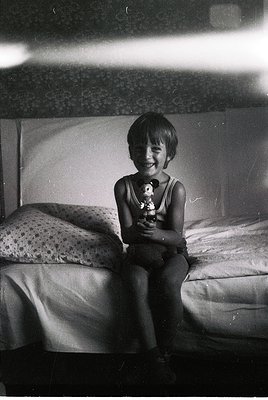 Vintage black-and-white photo of a smiling child in a sleeveless shirt holding a stuffed toy, seated on a bed with floral-pat...
