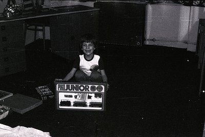 Child poses with a vintage **Junior OO gauge model train set** (1960s-70s) in a dimly lit indoor setting. Noticeable details:...