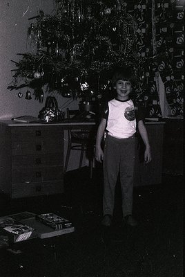 Vintage indoor holiday scene featuring a young child in 1970s-style clothing (short-sleeve shirt, pleated pants) standing bes...