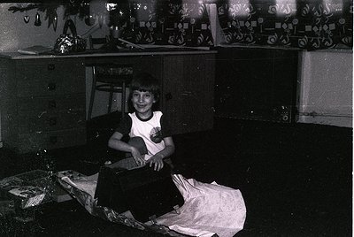 Black-and-white interior shot of a person seated on a bed in a modest, cluttered room. Decorative wallpaper with geometric pa...