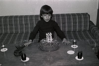 Young boy in 1970s-style sweater poses with birthday cake adorned with 11 lit candles on a dark wooden table. Plain checkered...