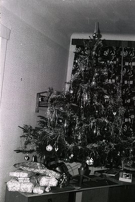 Mid-century Christmas tree adorned with tinsel, paper ornaments, and a star topper, placed beside wrapped gifts and a shelf o...