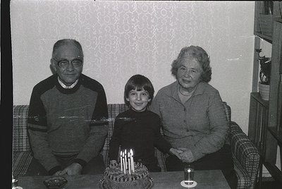 Black-and-white family portrait from the 1970s–1980s, featuring a young boy seated between two elderly adults on a patterned ...