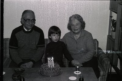 Family portrait featuring a grandfather, grandmother, and child seated at a table with a birthday cake adorned with lit candl...