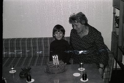 A tender 1970s family moment: a young boy and elderly woman pose beside a round birthday cake with lit candles. The woman wea...