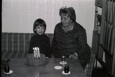 A black-and-white indoor portrait from the 1970s–1980s, featuring a grandmother and young boy at a birthday table. The boy, s...